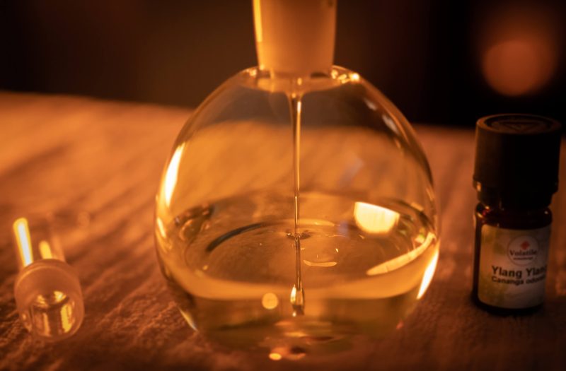 Een glazen fles massageolie en een flesje ylang-ylang op de massagetafel, in warm licht