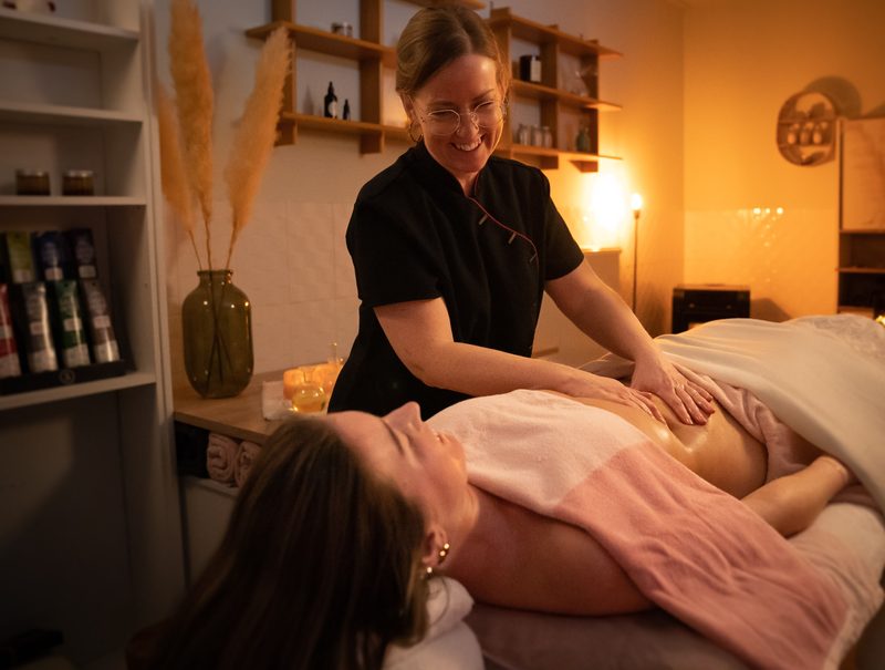 Lesley masseert lachend een cliënte op de massagetafel, in haar praktijkruimte bij warm licht