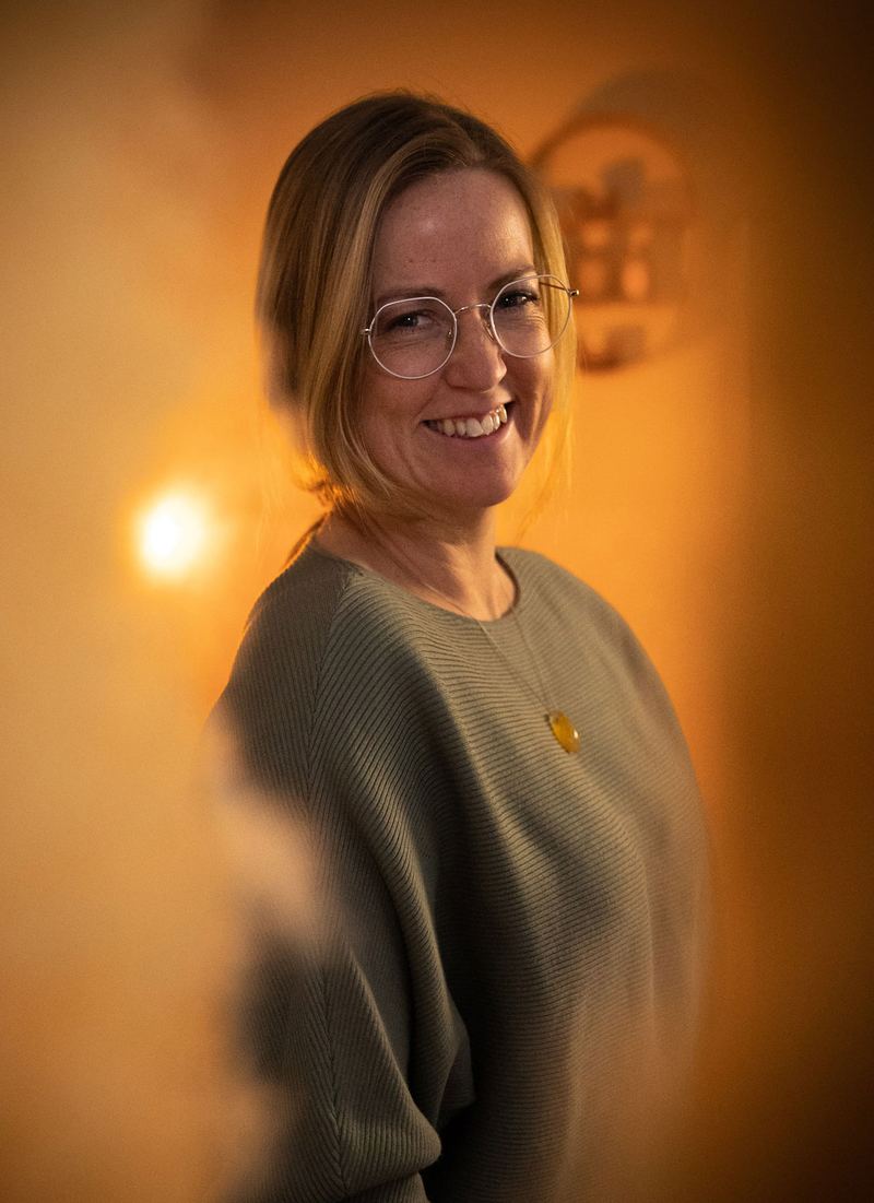 Lesley Lucas glimlacht warm over haar schouder, in een groene trui, bij kaarslicht